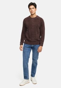 Jonge man die staat in een bruine gestructureerde trui, blauwe jeans en witte sneakers, tegen een effen lichte achtergrond.