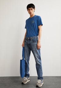 T-shirt in cotone blu con scollo rotondo, jeans azzurri chiari con zone sbiadite e sneakers grigie. Gli accessori includono una borsa tote blu.
