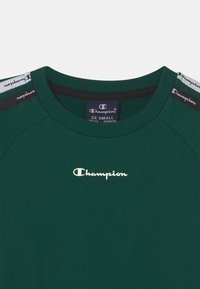 Zelené tričko s kulatým výstřihem, vyrobené z měkké tkaniny. Obsahuje bílé logo "Champion" a černobílé pruhy na ramenou.