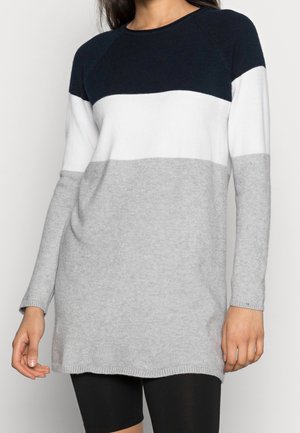 Vrouw draagt een lange mouw gebreide trui met horizontale kleurvlakken in marineblauw, wit en lichtgrijs, gecombineerd met zwarte korte broek.