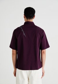 Kortermet skjorte i mørk lilla med rutemønster, med broderte detaljer på baksiden og en uformell kragedesign.