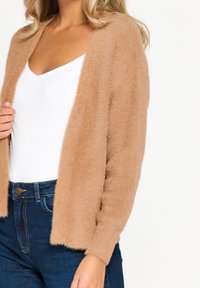 Cardigan de color marrón suave con una textura esponjosa, de frente abierto y mangas largas, llevado sobre una blusa blanca y combinado con jeans azul oscuro.