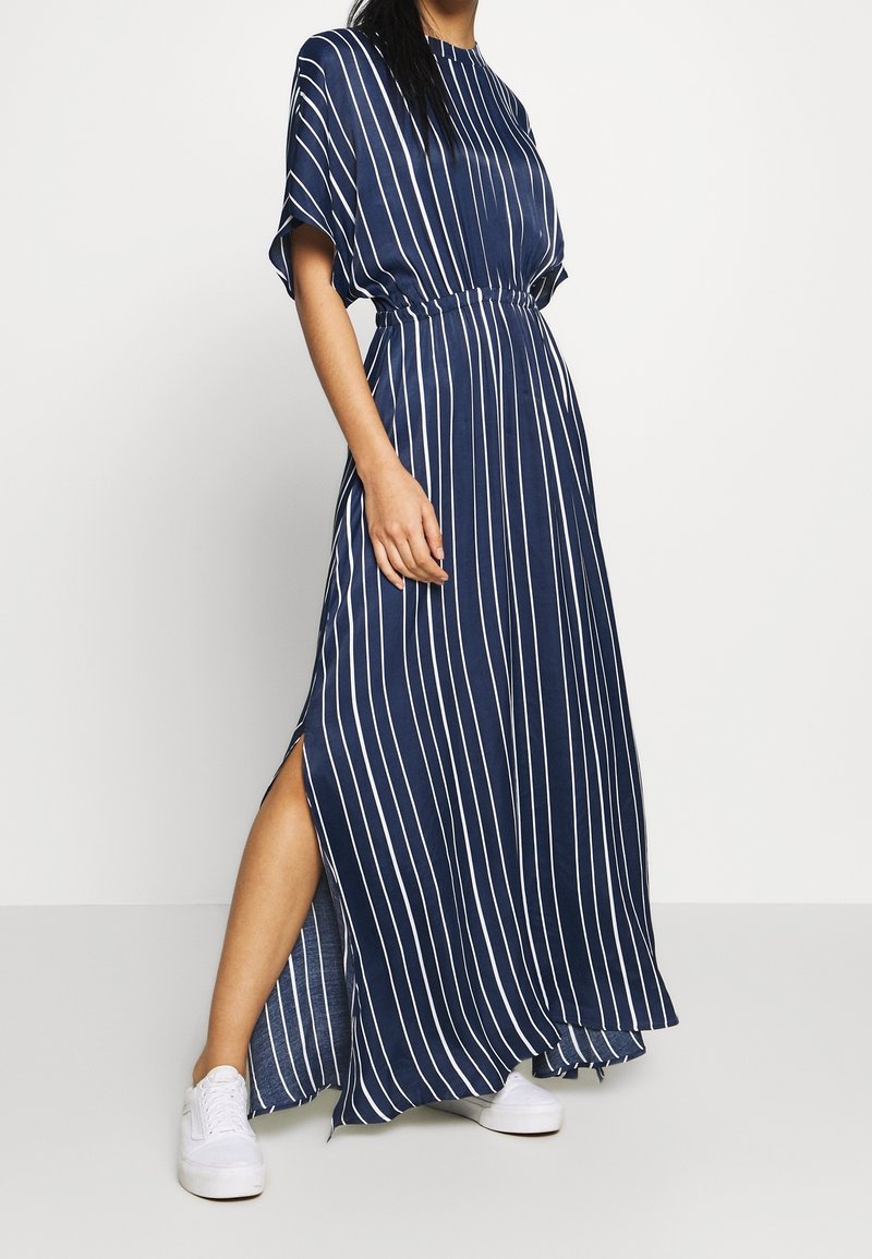 Femme portant une robe maxi à rayures verticales bleu marine et blanc avec une fente latérale et des baskets blanches, debout devant un fond uni.