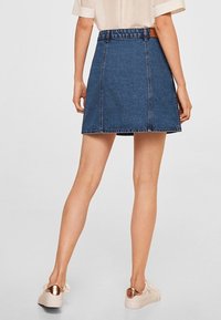Falda de mezclilla en azul oscuro, con una cintura ajustada y forma en A. Presenta costuras visibles y un dobladillo sin acabar. Combinada con zapatillas ligeras.