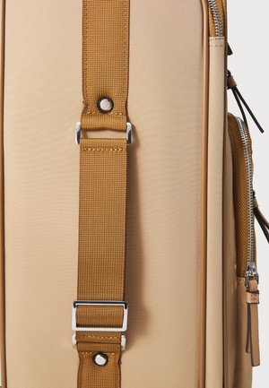 Beige utazótáska, amely textúrált külsővel, barna nylon pánttal ezüst csattal és oldalán zipzáros zsebbel rendelkezik.