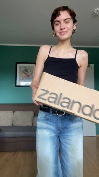 Smeđi kartonski kutija s natpisom "zalando" ispisanim sivo, drži osoba u crnoj majici i plavim trapericama, stojeći u sobi.