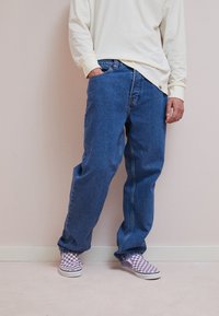 Persoon in losse blauwe jeans en paars-witte geruite instappers, staande tegen een lichte muur met één hand in de broekzak.