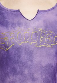 Fialové tričko s tónováním batikou, které má zlatý grafický textový design s nerovnými okraji. Hladký výstřih, měkká textilní struktura a krátké rukávy.