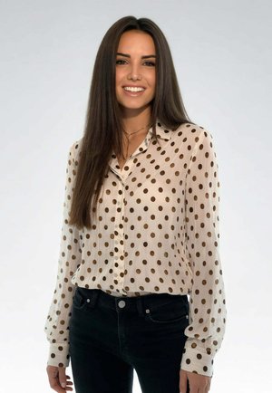 Giovane donna con lunghi capelli scuri che indossa una camicetta bianca con pois marroni e pantaloni neri, sorridente, in piedi contro uno sfondo chiaro.