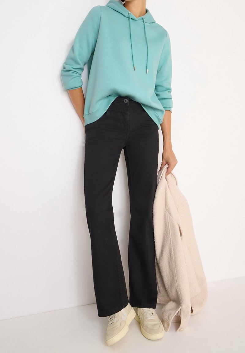 Personne portant un sweat à capuche bleu sarcelle, un pantalon noir et des baskets beiges, tenant une veste texturée beige clair dans une main sur un fond blanc.