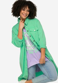 Camicetta oversize verde menta con bottoni a pressione, indossata sopra una t-shirt grafica viola chiaro con un design in paillettes, abbinata a jeans blu chiaro.