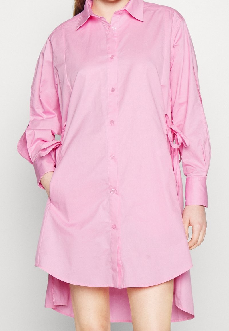 Robe chemise rose à boutons avec manches longues, poche avant et détails noués sur les manches. Fabriquée en tissu lisse avec une coupe décontractée.