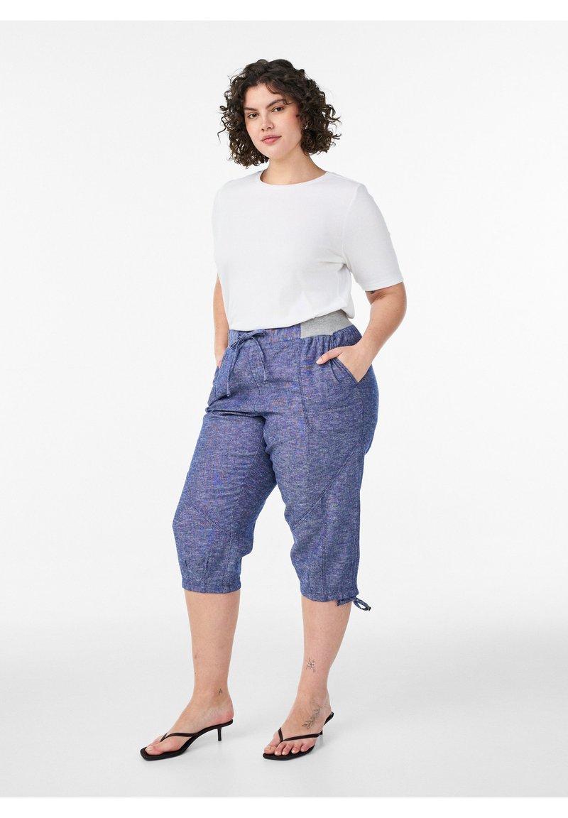 Femme aux cheveux bouclés portant un top blanc à manches courtes, un pantalon bleu court avec cordon et des sandales noires à talons, debout les mains dans les poches.