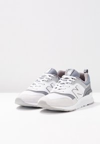 Ett par grå och vita New Balance-sneakers med snörning, nät- och mockadetaljer, placerade på en vit yta mot en enfärgad bakgrund.