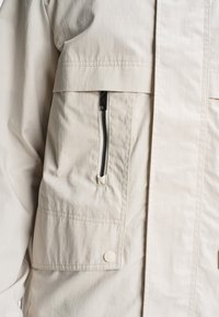 Veste beige avec poche zippée verticale noire et poche à rabat avec bouton-pression sur la partie avant du torse.