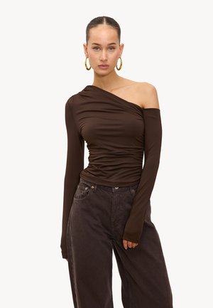 Femme portant un haut à une épaule marron foncé à manches longues et un jean ample foncé, avec des boucles d'oreilles en or et les cheveux coiffés en arrière.