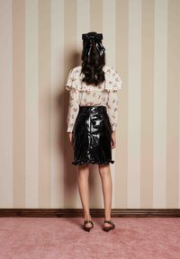 Saia de couro patente preta com bainha rodada, combinada com uma blusa de manga longa transparente com padrão floral e uma faixa de cabelo preta com laço.