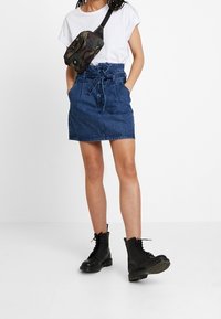 Jeansrock mit hoher Taille, Bindegürtel und seitlichen Taschen, kombiniert mit einem weißen T-Shirt und schwarzen Schnürstiefeln.