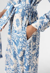 Abito floreale blu e bianco con maniche lunghe, vita con cintura, tasche e tessuto decorato con grandi fiori e foglie.