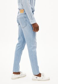 Jeans en denim bleu clair avec une coupe droite, arborant une patch en cuir sur la ceinture arrière, portés avec des bateaux blancs.