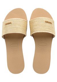 Tasaplantut sandaalit, joissa on leveä, teksturoitu beige hihna ja kumipohja. Hihnassa on kultainen logo. Pohjassa on pisteellinen pinta paremman pidon saavuttamiseksi.