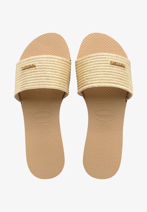 Sandale plate cu o bandă lată, texturată, de culoare bej și o talpă din cauciuc. Prezentă un logo aurit pe bandă. Talpa are o suprafață punctată pentru aderență.