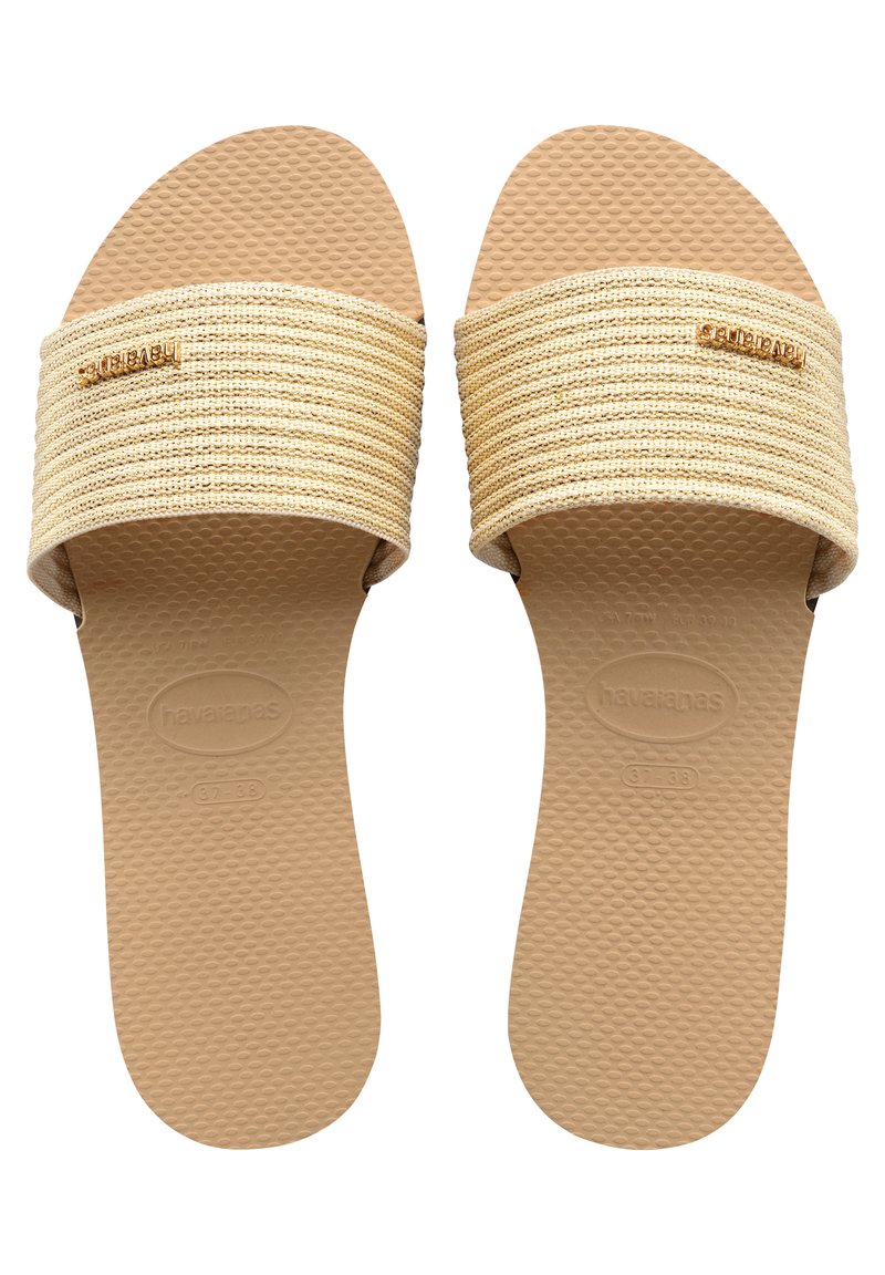Tasaplantut sandaalit, joissa on leveä, teksturoitu beige hihna ja kumipohja. Hihnassa on kultainen logo. Pohjassa on pisteellinen pinta paremman pidon saavuttamiseksi.