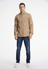 Beige overhemd met lange mouwen en knopen, voorzien van een zak; gecombineerd met donkerblauwe jeans en witte sneakers. De stof lijkt soepel met een lichte textuur.