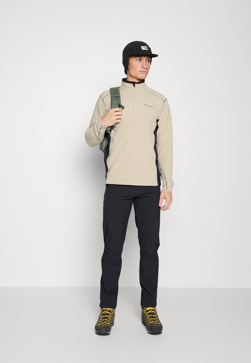 Beige Fleecejacke mit Reißverschluss und schwarzen Seitenpaneelen, kombiniert mit schwarzen Hosen und Wanderschuhen. Die Person trägt eine schwarze Mütze und hat einen grünen Rucksack dabei.