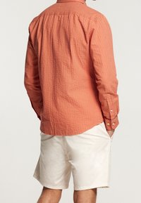 Red and white striped long-sleeve shirt with a back pleat and buttoned cuffs, paired with beige shorts. Textured, casual fit.