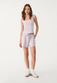 Ljubičasti crop top u kombinaciji s ljubičastim šortsevima s povlačenjem. Ima glatku tkaninu, bočne džepove i srebrne tenisice s bijelim i krem akcentima.