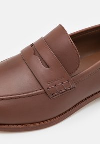 Mocassins en cuir marron avec une texture lisse, dotés d'une sangle décorative et d'accents de couture. Logo embossé sur le côté.
