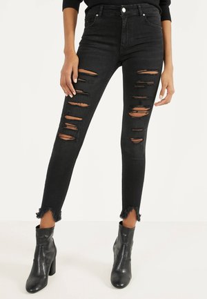 Musta revityt skinny-farkut, joissa on repaleiset reunat, yhdistettynä mustiin korollisiin nilkkureihin, näkyvissä vyötäröstä jalkoihin vaalealla taustalla.