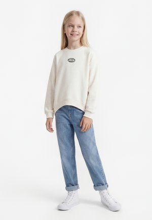 Junges Mädchen mit langen blonden Haaren, das einen cremefarbenen Sweatshirt, hellblaue hochgekrempelte Jeans und weiße High-Top-Sneaker trägt, steht und lächelt.