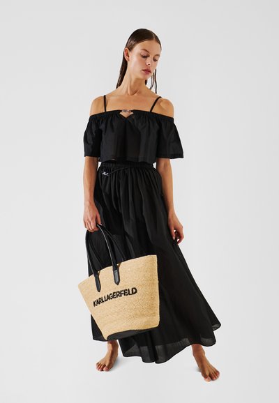 Haut noir à épaules dénudées avec des manches à volants, associé à une jupe maxi noire et un sac fourre-tout en paille avec des poignées noires et un logo.