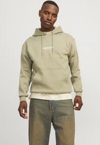 Olijfgroen hoodie met een voorzak, verstelbaar koord en witte tekstbranding. Gelaagd over een lichtgepatroniseerd shirt, gecombineerd met blauwe jeans.
