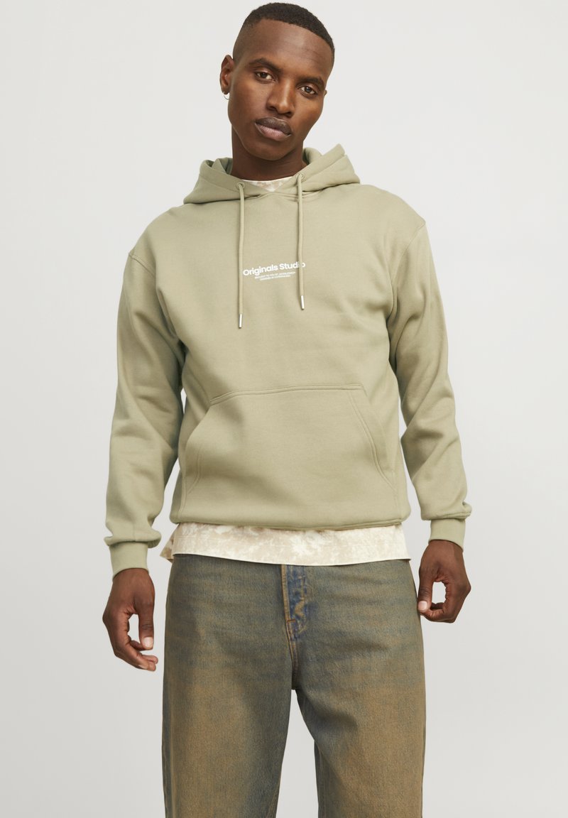 Olijfgroen hoodie met een voorzak, verstelbaar koord en witte tekstbranding. Gelaagd over een lichtgepatroniseerd shirt, gecombineerd met blauwe jeans.