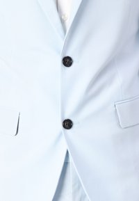 Világoskék blazer szövetből, két elülső zsebbel, három fekete gombbal és egy nyílással a hátán.
