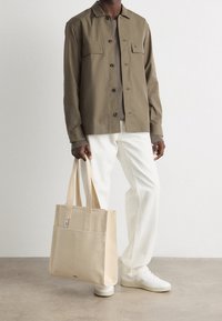 Béžová plátěná taška na rameno s dvojitými držadly, obdélníkovým tvarem, přední kapsou a jemnými detaily, spárovaná se zelenošedou košilí a světle zbarvenými kalhotami.