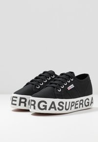 Black canvas sneakers with a textured upper, white rubber platform sole, and bold black lettering "SUPERGA" on the side. Classic lace-up design.