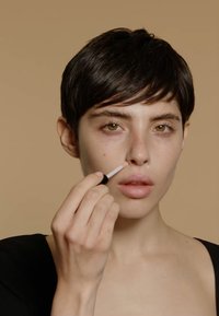 Aplicador de labios con punta afilada sostenido por un modelo, aplicando un producto beige claro en el labio superior. Cabello corto y oscuro con fondo neutro.