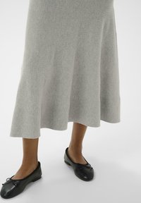 Jambes inférieures portant une jupe midi gris clair et des ballerines en cuir noir avec des petits nœuds sur un fond clair 
