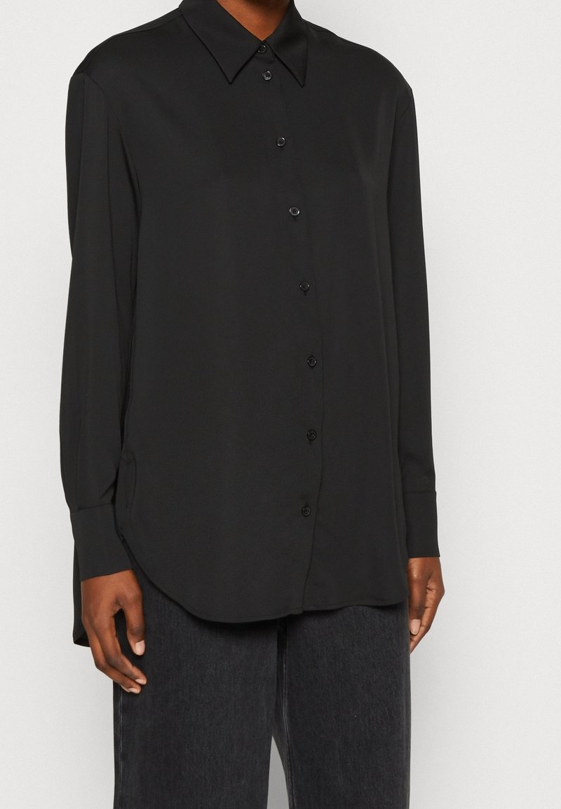 Personne portant une chemise noire ample à boutons, manches longues et col pointu, associée à un pantalon gris foncé.