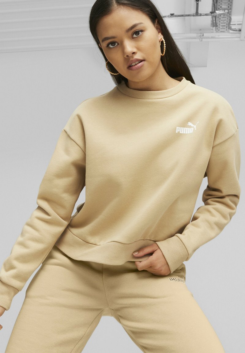 Felpa beige con polsini a costine, scollatura rotonda e piccolo logo bianco. Abbinata a pantaloni beige coordinati, realizzati in materiale morbido e testurizzato.