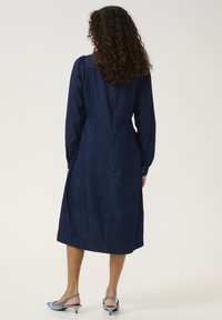 Mörk denim långärmad klänning med skräddarsydd passform, som har en bakseam, knäppte manschetter och en nederkant under knäna. Bärs med ljusblå klackar.