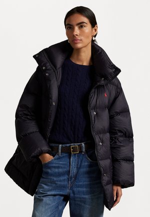 Chaqueta tipo puffer negra con cuello alto, botones a presión y un logotipo rojo. Combinada con un suéter de punto en color azul marino y jeans azules.