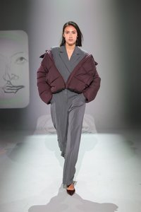 Grynasis pūkinis švarkas tamsiai burgundiškai raudonos spalvos, uždėtas ant pilkos juostuotos kostiumo. Modelis vilki rudas geometrines aukštakulnes. Paprasta fono kompozicija su švelniu apšvietimu.