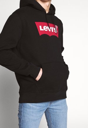 Osoba w czarnej bluzie z kapturem Levi's z czerwono-białym logo, z rękami w przedniej kieszeni, ubrana w jasnoniebieskie dżinsy.