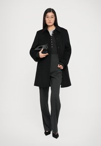 Long black coat over a gray fitted top and pinstriped trousers, holding a black clutch. Features include a structured collar and tailored fit.