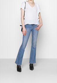 Top bianco a maniche corte con scollo a V e bordi arricciati, abbinato a jeans svasati blu chiaro e stivali neri alla caviglia. Completo semplice e casual.
